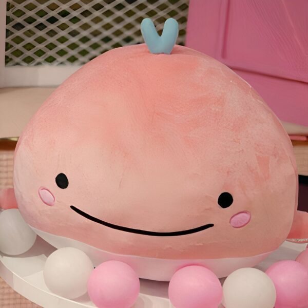 Une baleine rose en peluche avec un sourire et un petit nœud bleu sur la tête, entourée de boules roses et blanches.