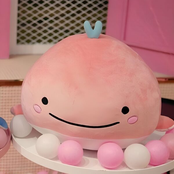 Une baleine rose en peluche avec un petit sourire et une nageoire bleue sur le dos, posée sur un lit de boules roses et blanches.