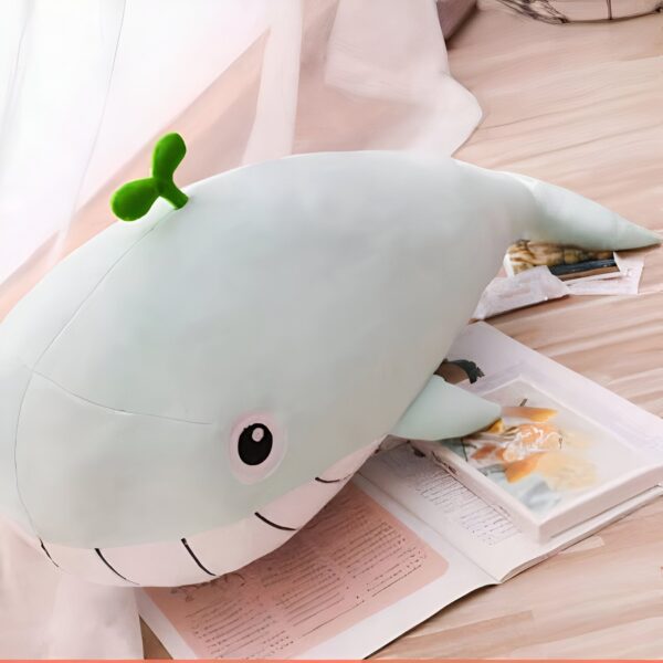 Une peluche de baleine verte avec un petit brin d'herbe sur la tête, posée sur un livre.