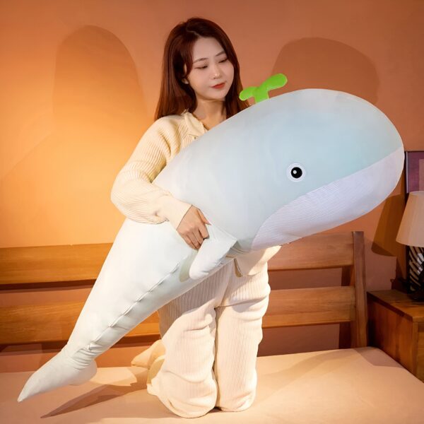 Une jeune femme souriante tient une grande peluche baleine bleue sur un lit. La baleine a une petite plante verte sur la tête.