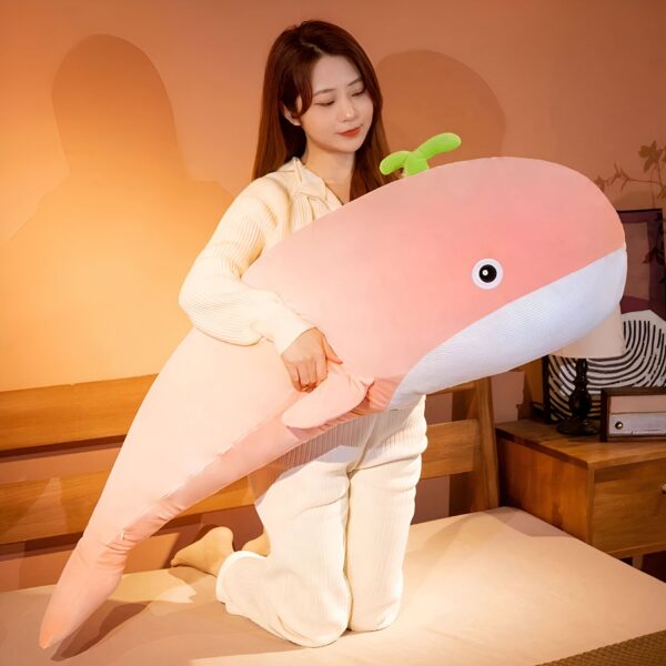 peluche-baleine-geante-pastel-xxl-cadeau-enfant-adulte-2a-3 Une jeune femme souriante tient une grande peluche rose en forme de baleine avec une petite plante verte sur la tête.