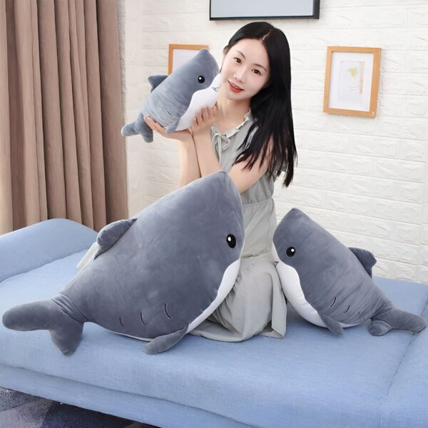 Une jeune femme est assise sur un canapé avec trois peluches en forme de requin. Elle tient un petit requin dans ses bras et les deux autres sont sur le canapé.