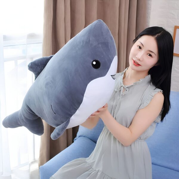 Une jeune femme souriante tenant un grand coussin en forme de requin gris.