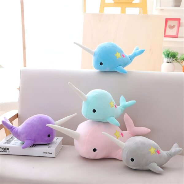 peluche-baleine-licorne-bleue-avec-etoiles-2a Cinq peluches narvals en bleu clair, bleu foncé, violet, rose et gris, disposées sur un canapé blanc. Chaque peluche a une petite étoile brodée sur le dos.