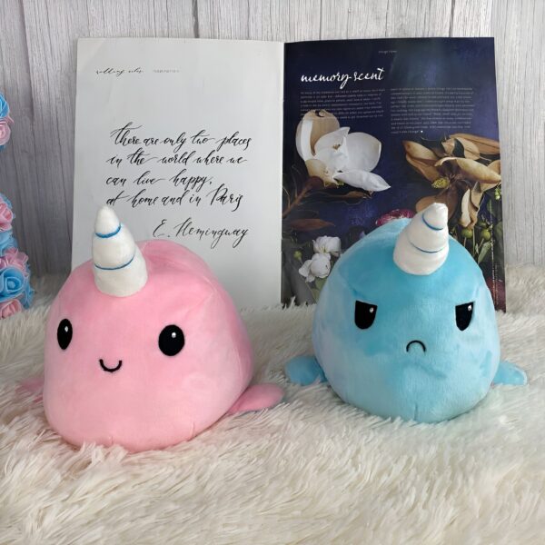Deux peluches en forme de narval, l'une rose avec un sourire et l'autre bleue avec une expression triste. Les peluches sont posées sur une surface en fourrure blanche.