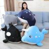 Une femme souriante est assise sur un canapé avec quatre gros animaux en peluche. Il y a une raie manta, un requin, une orque et une narval.