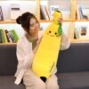 Une jeune femme souriante tient dans ses bras un oreiller en forme de banane jaune avec un visage souriant et l'inscription "I love banana". L'oreiller est placé sur un canapé beige.