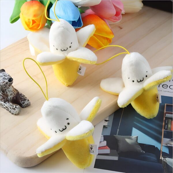 Trois peluches en forme de banane jaunes et blanches avec des visages souriants. Les peluches sont posées sur une surface en bois avec un bouquet de tulipes en arrière-plan.