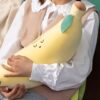 peluche-banane-jouet-doux-et-confortable-pour-enfants-2a Une femme tient un coussin en forme de banane jaune avec un visage souriant et une feuille verte.