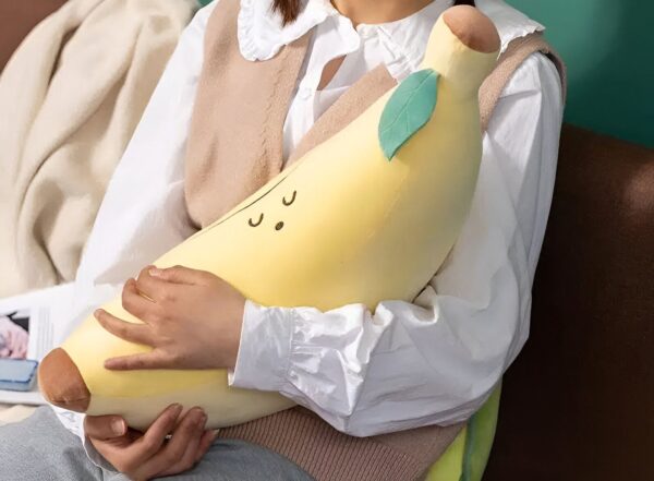 peluche-banane-jouet-doux-et-confortable-pour-enfants-2a Une femme tient un coussin en forme de banane jaune avec un visage souriant et une feuille verte.