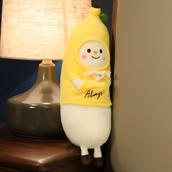 peluche-banane-kawaii-oreiller-doux-et-moelleux-3a Un coussin en forme de banane souriant avec un sweat-shirt jaune et l'inscription "Always" sur un lit avec une lampe de chevet.