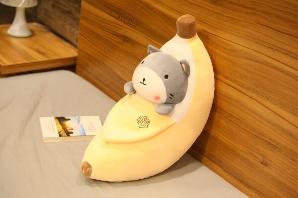 Un coussin en forme de banane avec un chat gris à l'intérieur, sur un lit blanc.