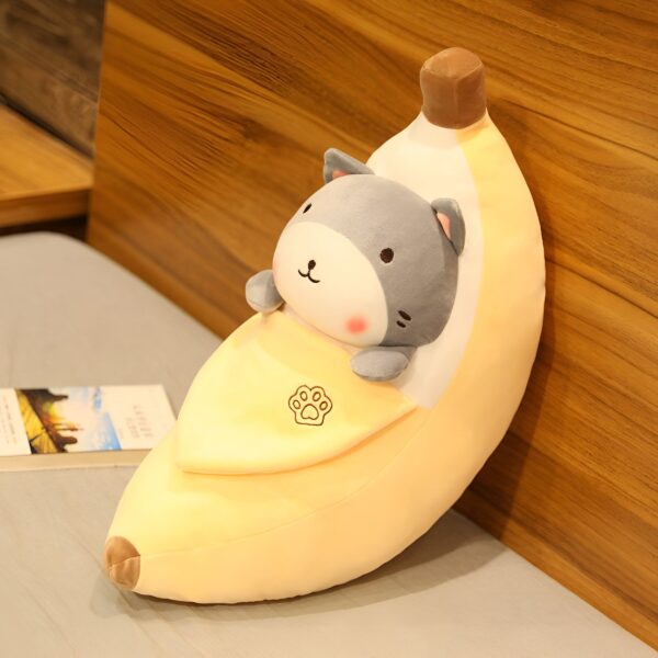 Un coussin en forme de banane avec un chat gris à l'intérieur, sur un lit blanc.