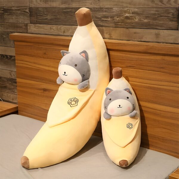 Deux peluches en forme de banane avec un chat gris à l'intérieur, l'une plus grande que l'autre. Les peluches sont posées sur un lit.