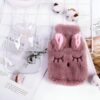 Bouillotte en forme de lapin en fourrure rose avec des oreilles roses et des yeux brodés. La bouillotte est posée sur un fond blanc avec des fleurs blanches.