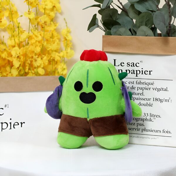 peluche-brawl-stars-spike-et-leo-pack-de-2-2a-2 Peluche en forme de cactus vert avec un visage souriant et des shorts bruns, tenant deux sacs violets. Il est assis sur une table blanche.