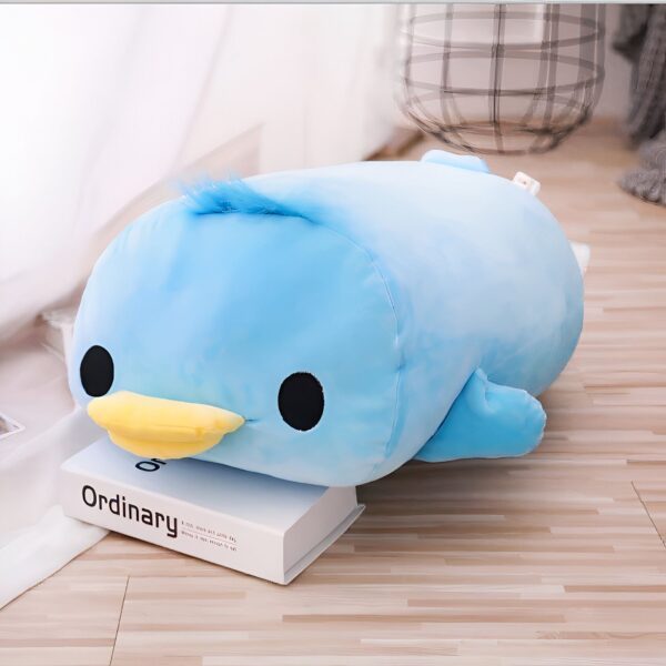 Une peluche bleue en forme de canard avec un bec jaune et des yeux noirs, allongée sur un livre sur un plancher en bois.