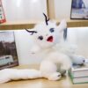 Peluche blanche avec des cornes noires, des yeux bleus et une bouche rose, assis sur une table en bois avec un livre à côté.