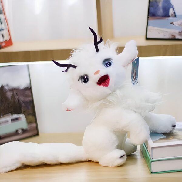 Peluche blanche avec des cornes noires, des yeux bleus et une bouche rose, assis sur une table en bois avec un livre à côté.