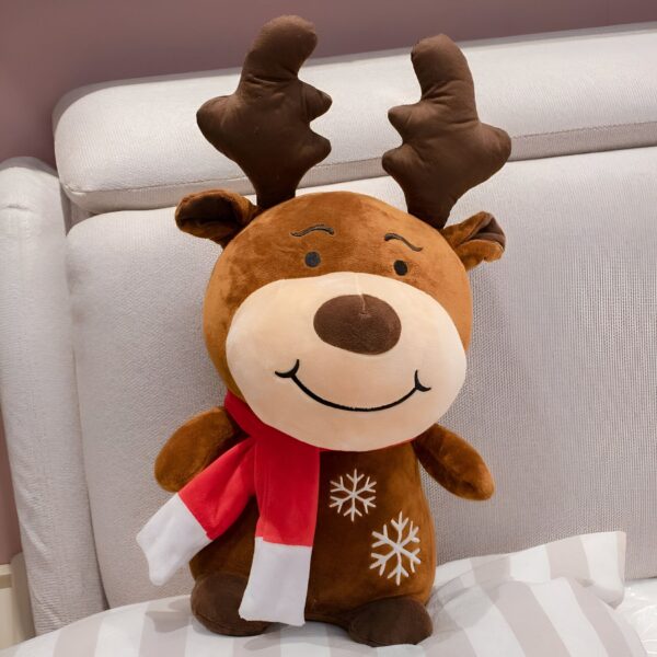 peluche-cerf-noel-jouet-doux-pour-enfants-1a-2 Un renne en peluche brun portant une écharpe rouge et blanche avec des flocons de neige brodés sur son corps, assis sur un canapé beige.