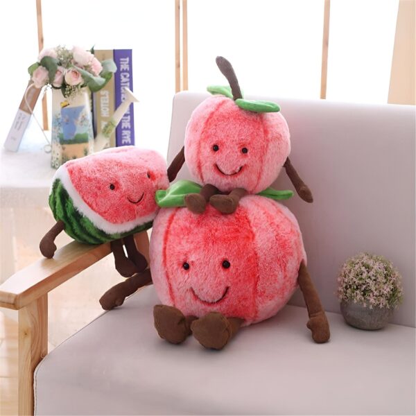 Trois peluches en forme de fruits souriants : une pastèque et deux pommes, assises sur un canapé blanc.