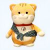 Une peluche de chat jaune portant un foulard vert avec des motifs. La peluche a un médaillon argenté autour du cou.