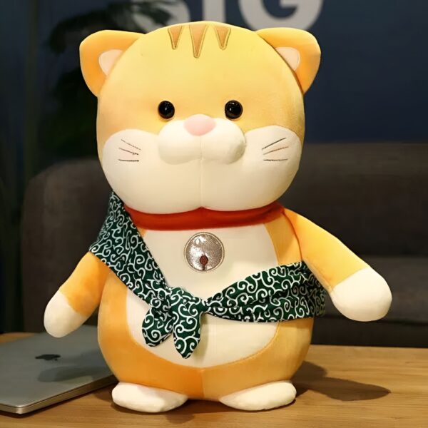 Peluche de chat jaune avec une écharpe verte et un collier rouge, assis sur une table en bois.