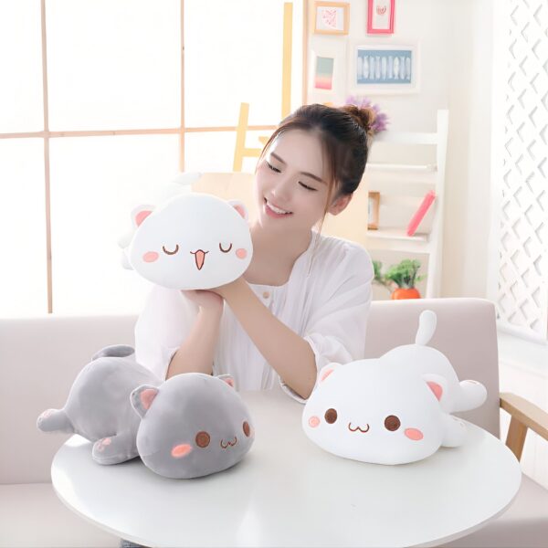 peluche-chat-blanc-kawaii-mignon-et-calin-ideal-pour-les-enfants-2a-2 Une jeune femme sourit en tenant un coussin en forme de chat blanc. Deux autres coussins en forme de chat, l'un blanc et l'autre gris, sont sur la table.