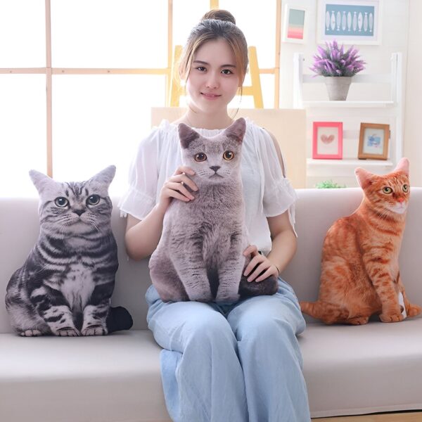 Une jeune femme assise sur un canapé avec trois coussins en forme de chat : un chat gris, un chat noir et blanc et un chat orange.