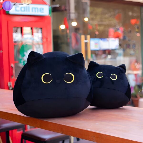 Deux coussins en forme de chat noir avec de grands yeux jaunes, posés sur une table en bois.