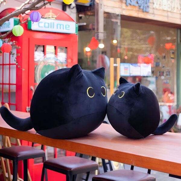 Deux peluches de chat noir rondes sur une table en bois devant une cabine téléphonique rouge. Les peluches ont de grands yeux jaunes. Les peluches sont un excellent moyen de décorer une chambre ou un salon.