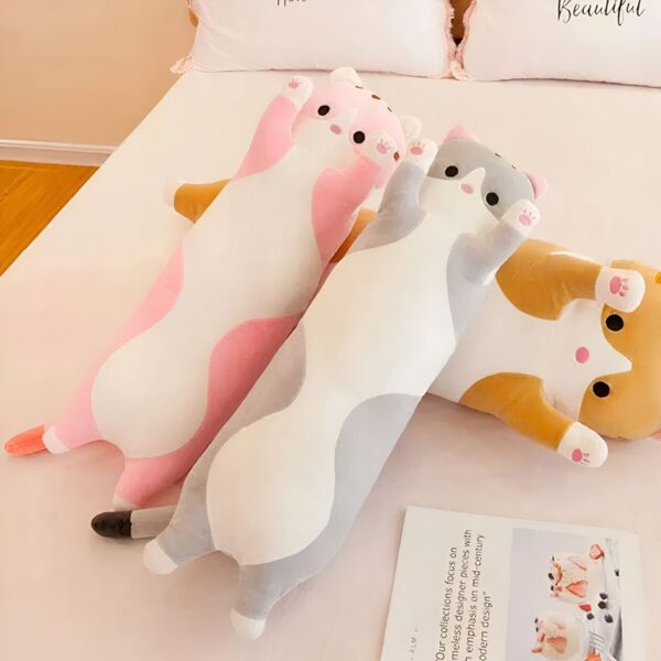 Trois coussins en forme de chat, un rose, un gris et un orange, posés sur un lit.
