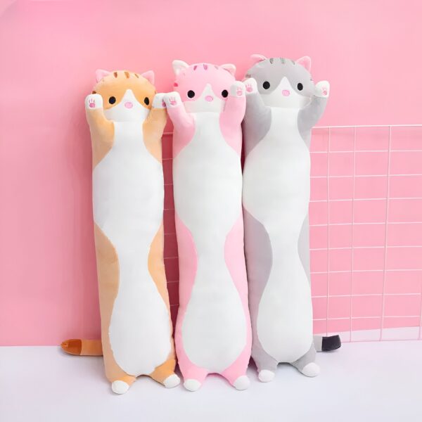 Trois coussins en forme de chat, un orange, un rose et un gris, posés devant un mur rose.