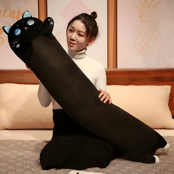 Une femme souriante est assise sur un lit et tient un oreiller en forme de chat noir. L'oreiller est long et doux.