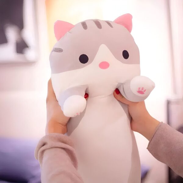 Une peluche de chat gris et blanc avec des détails roses est tenue par deux mains. La peluche a de grands yeux noirs et un nez rose.