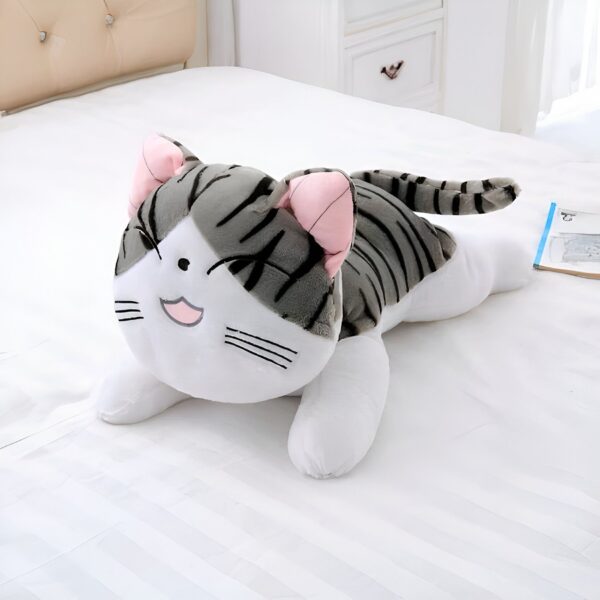 Peluche chat gris et blanc allongé sur un lit blanc. Il a de grandes oreilles roses et sourit.