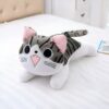 Une peluche de chat gris et blanc avec des yeux noirs et une bouche rose, allongée sur un lit blanc.