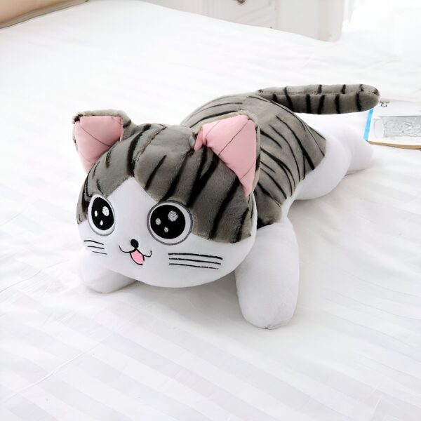 Une peluche de chat gris et blanc avec des yeux noirs et une langue rose, allongé sur un lit blanc.