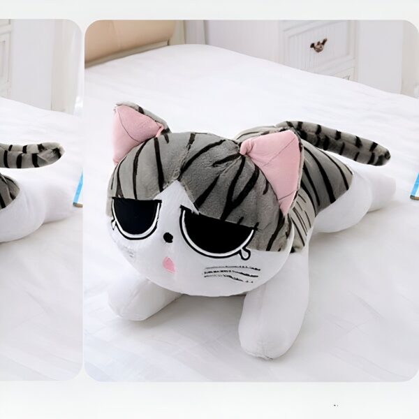 Peluche chat gris et blanc allongé sur un lit blanc. Le chat a de grands yeux noirs et des oreilles roses.