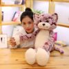 Une jeune femme souriante tient un grand chat en peluche rose et blanc. Le chat a de grands yeux noirs et un nez rose.