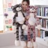 Une jeune femme tient deux grandes peluches de chat, l'une grise et l'autre rose. Les chats ont de longues jambes et de grandes têtes.
