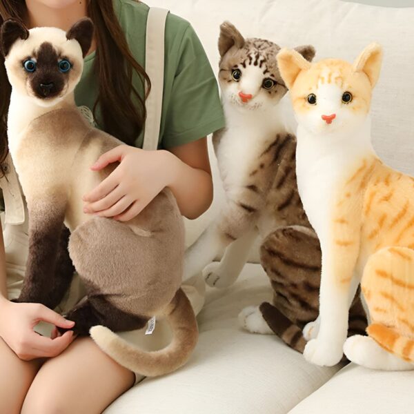 peluche-chat-siamois-gris-jouet-doux-et-calin-lavable-en-machine-2a Trois peluches de chats réalistes : un chat siamois, un chat tabby et un chat orange. Les chats sont assis sur un canapé blanc.