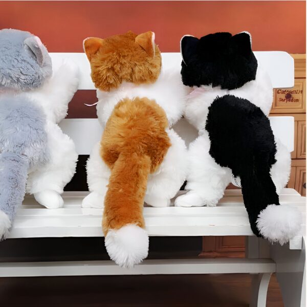 Trois peluches de chat assis sur un banc blanc. Le chat à gauche est gris et blanc, le chat au milieu est orange et blanc, et le chat à droite est noir et blanc.