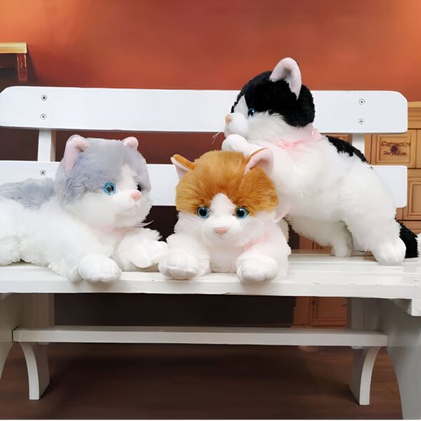 Trois peluches de chat assises sur un banc en bois blanc. Le chat à gauche est gris et blanc, celui du milieu est orange et blanc et celui de droite est noir et blanc.