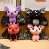 Quatre peluches de chauve-souris avec des ailes noires. Une chauve-souris violette avec une étoile à cinq branches, une chauve-souris rouge avec une étoile à cinq branches, une chauve-souris grise avec une lune et une chauve-souris rose.