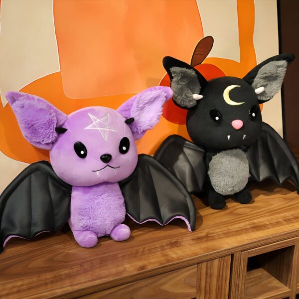 Deux chauves-souris en peluche, l'une violette avec une étoile à cinq branches sur le front et l'autre noire avec une lune sur le front.