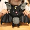 Une peluche noire en forme de chauve-souris avec des ailes en cuir noir, tenant une lune dorée sur la tête.