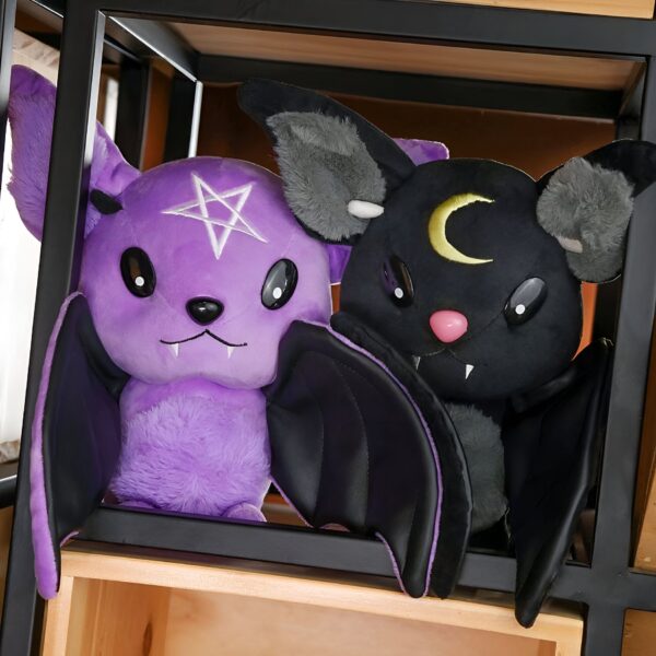 Deux chauves-souris en peluche, l'une violette avec une étoile à cinq branches et l'autre noire avec une lune, sont placées sur une étagère en bois.