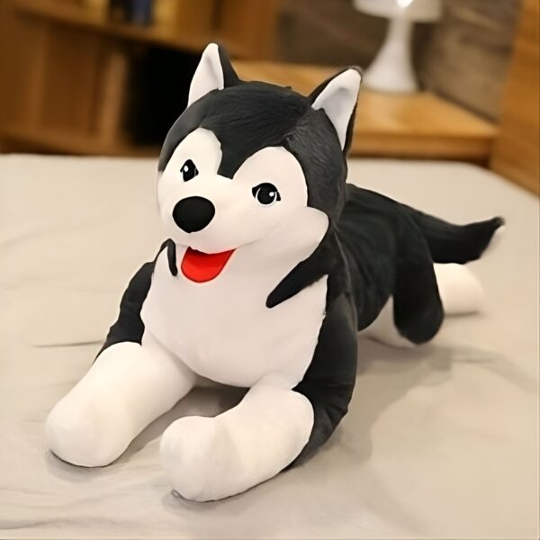 Une peluche de chien husky noir et blanc couché sur un lit. Le husky a une langue rouge et semble heureux.