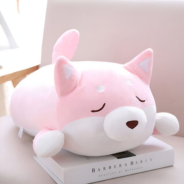 peluche-chien-kawaii-rose-peluche-doudou-1a Un coussin en forme de chien rose avec des yeux fermés, couché sur un livre.